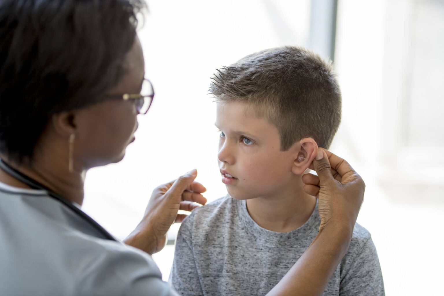 A little boy is sitting on an examining table in a doctors office - a healthcare professional is implanting a hearing aid for the child.