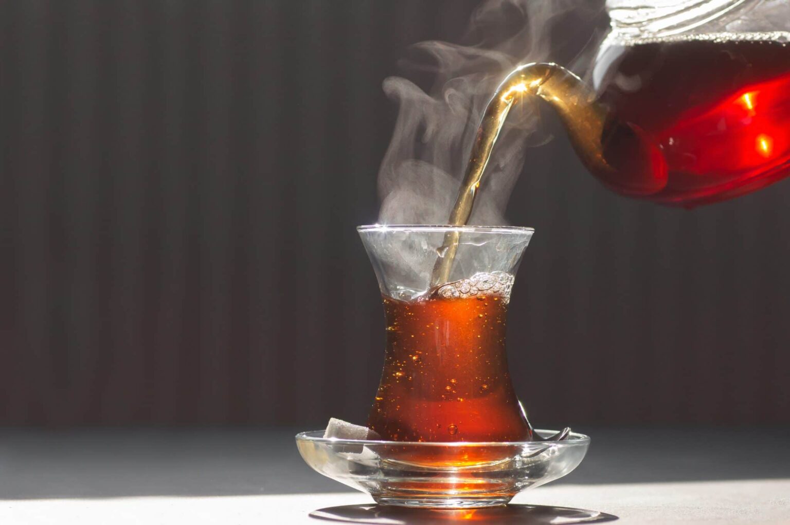 Glass cup of brewed black turkish tea pouring from teapot, traditional hot drink concept