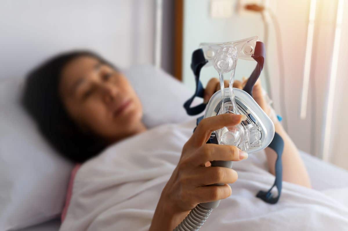 Sleep-Apnea-treatment Woman in a hospital clinical setting is laying on a hospital bed, she is blurry and the focus of the image is on the half-mask for a cpap that she is holding up in front of herself.